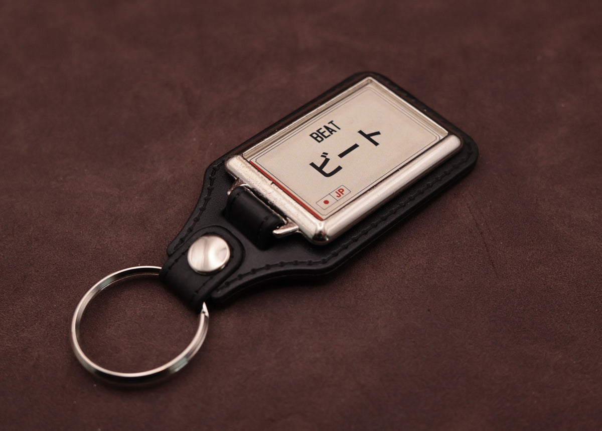 Honda Beat Katakana keyring angled with the split ring at lower left, showing the cream insert and black leatherette fob