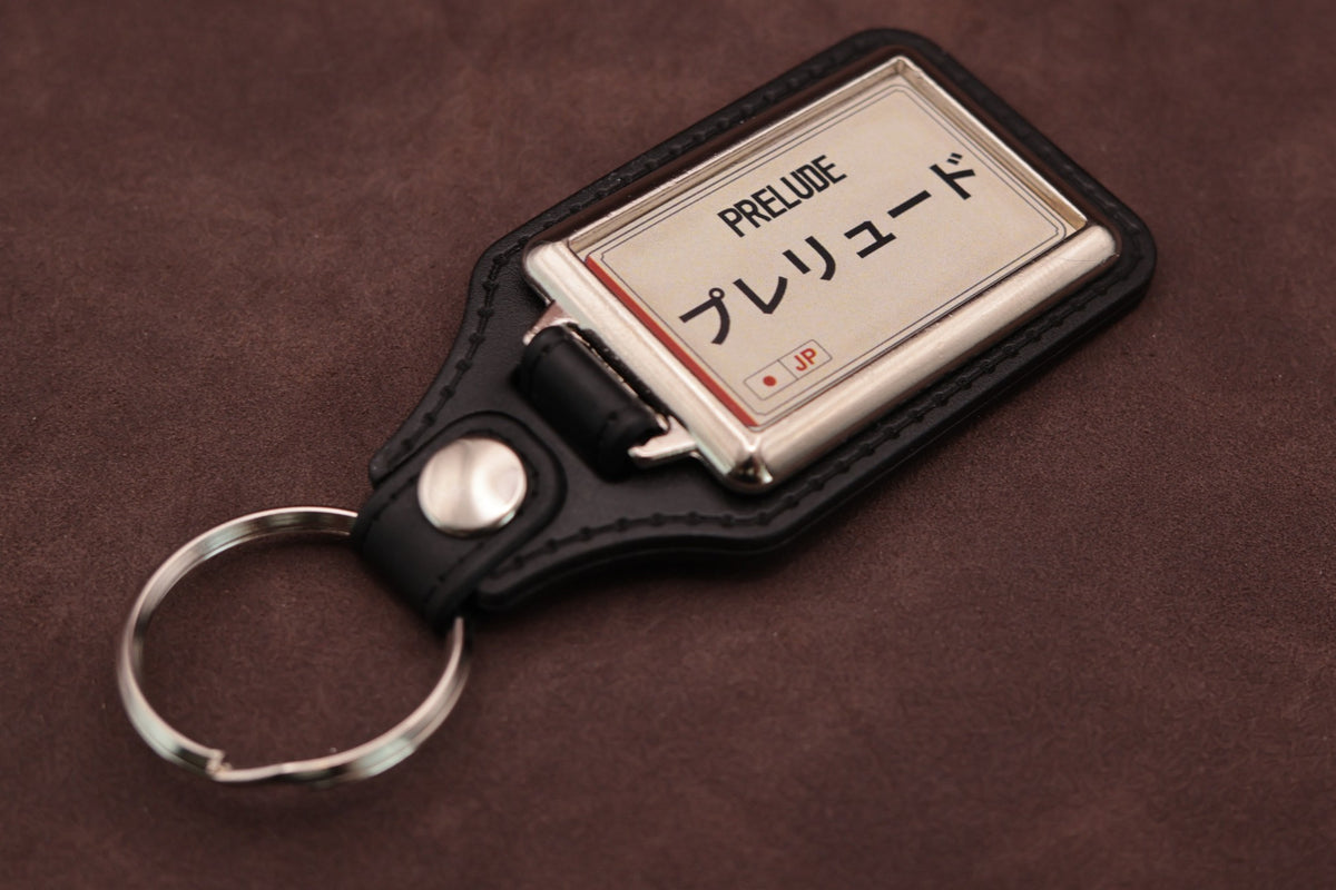 Honda Prelude Katakana keyring angled with the split ring at lower left, showing the cream insert and black leatherette fob