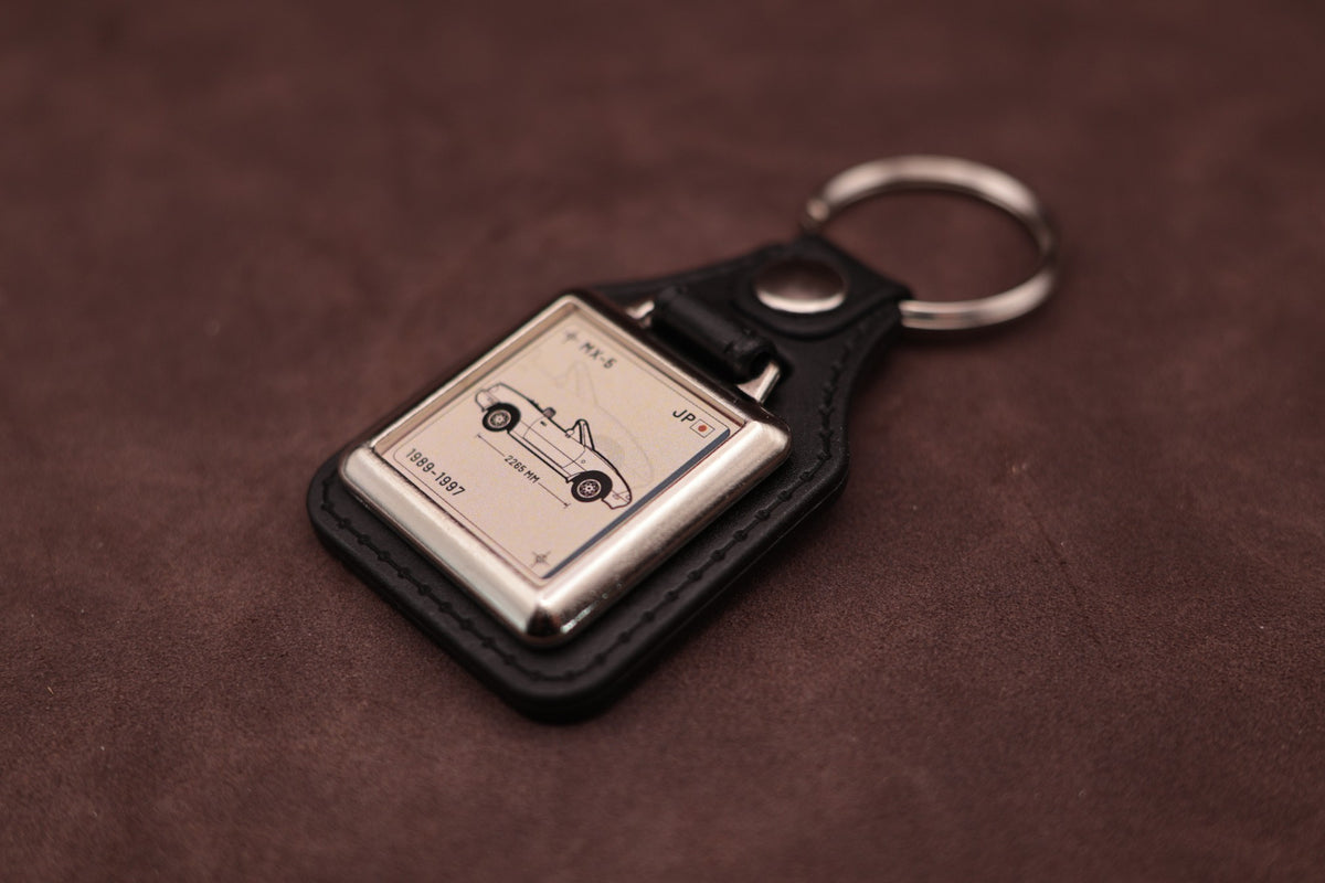 Mazda MX-5 NA Register Series keyring angled with the split ring at upper right and ivory vehicle artwork visible