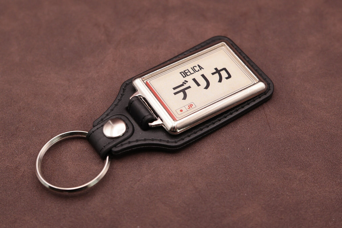Mitsubishi Delica Katakana keyring angled with the split ring at lower left, showing the cream insert and black leatherette fob