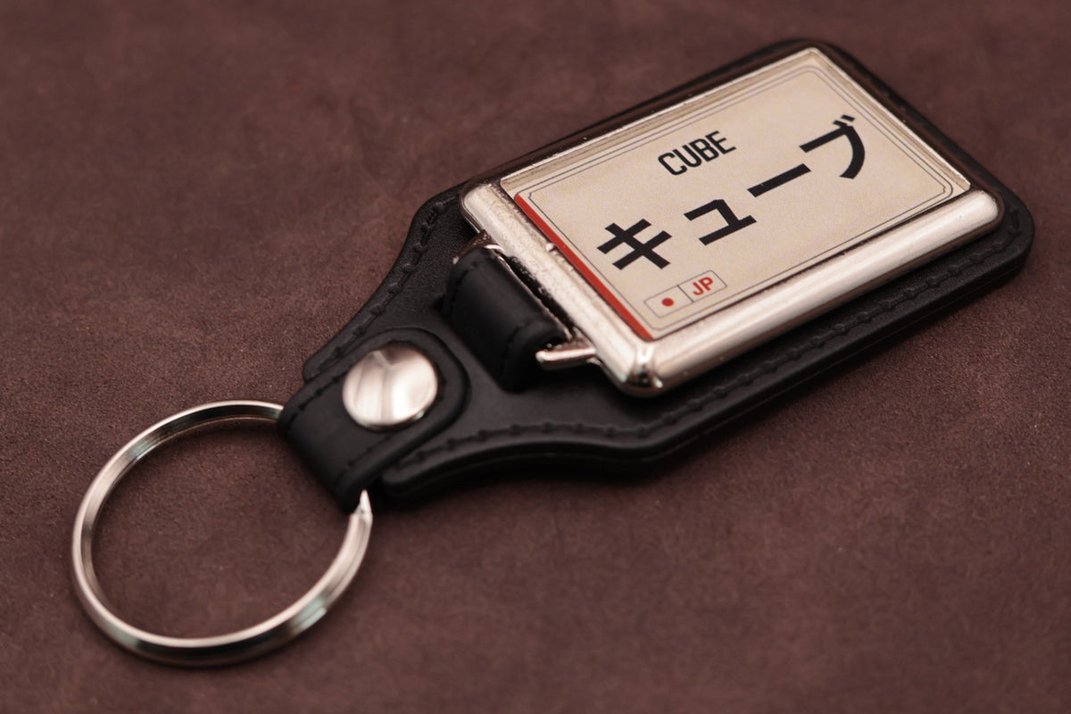 Nissan Cube Katakana keyring angled with the split ring at lower left, showing the cream insert and black leatherette fob
