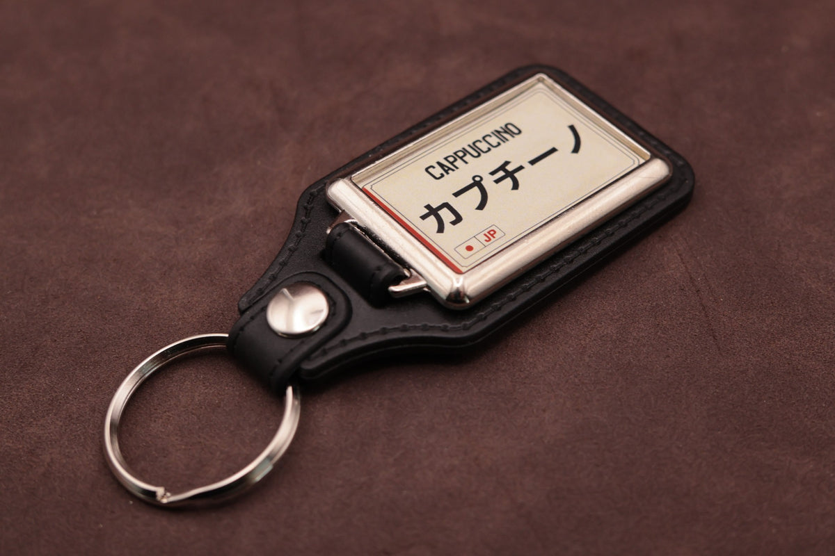 Suzuki Cappuccino Katakana keyring angled with the split ring at lower left, showing the cream insert and black leatherette fob