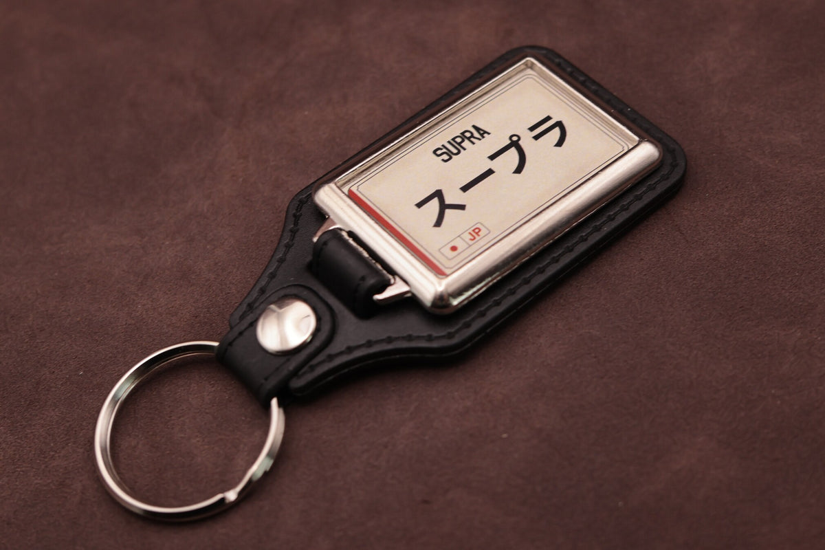 Toyota Supra Katakana keyring angled with the split ring at lower left, showing the cream insert and black leatherette fob